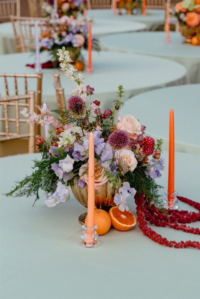 A colorful floral centerpiece alongside orange tapered candles and citrus are photographed at a Birmingham wedding reception at The Sage