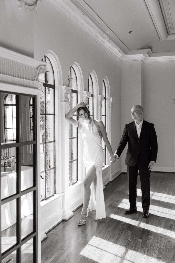 A black and white portrait shows a bride leaning against a window while holding hands with her husband at their reception held at The Florentine Birmingham