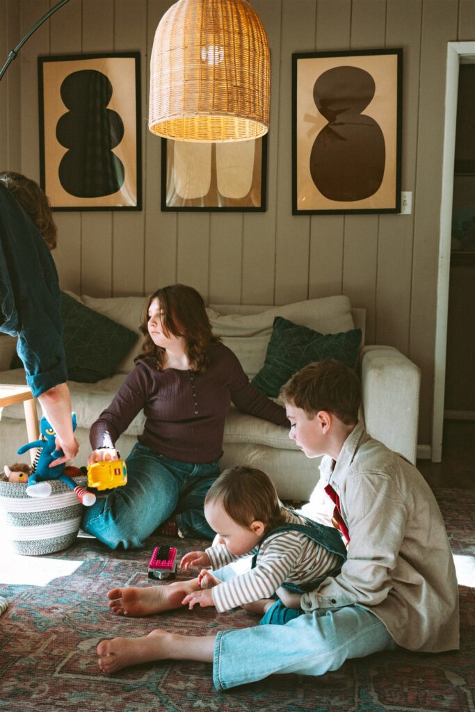A candid image depicts older siblings playing in their living room floor with their toddler brother during an in-home session with a family photographer birmingham al