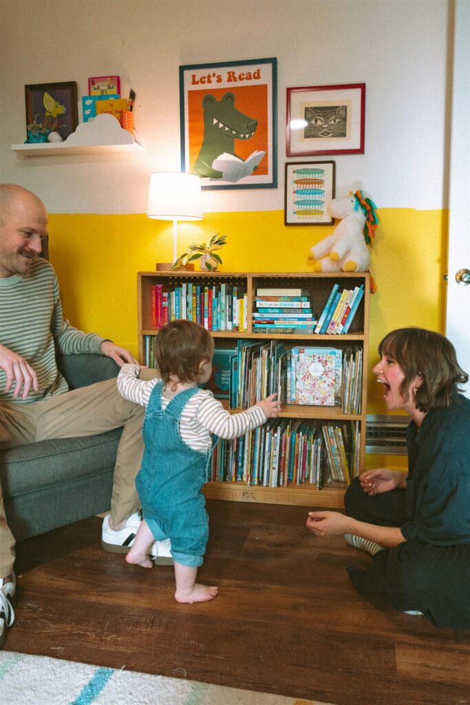 A toddler browses his bookshelf in his yellow & white nursery as his parents look on during family portraits birmingham