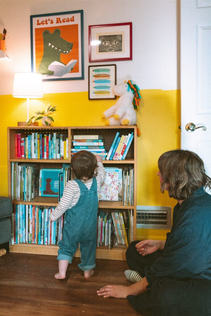 A toddler boy picks a book from the shelf in his mustard yellow nursery as his mother watches during a photoshoot in their home with an alabama family photographer