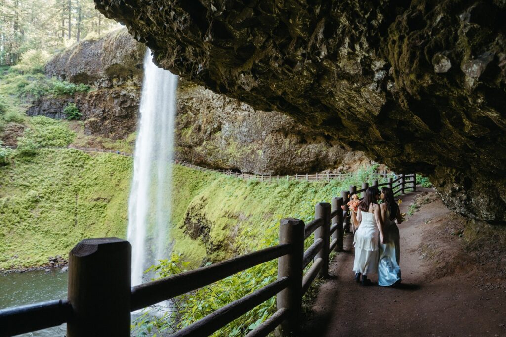 A couple walks beneath a waterfall in their wedding gowns while eloping in Oregon