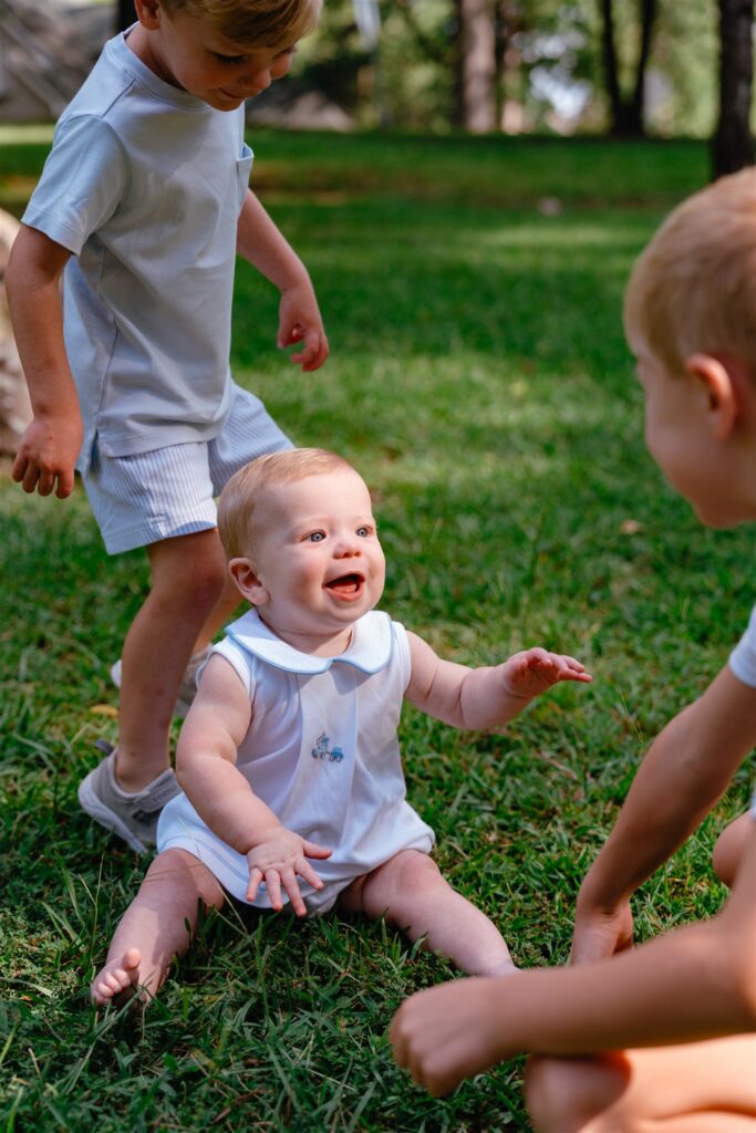 A baby plays in the grass at a local park while two older siblings look on in a candid photo taken during family photography Birmingham. 