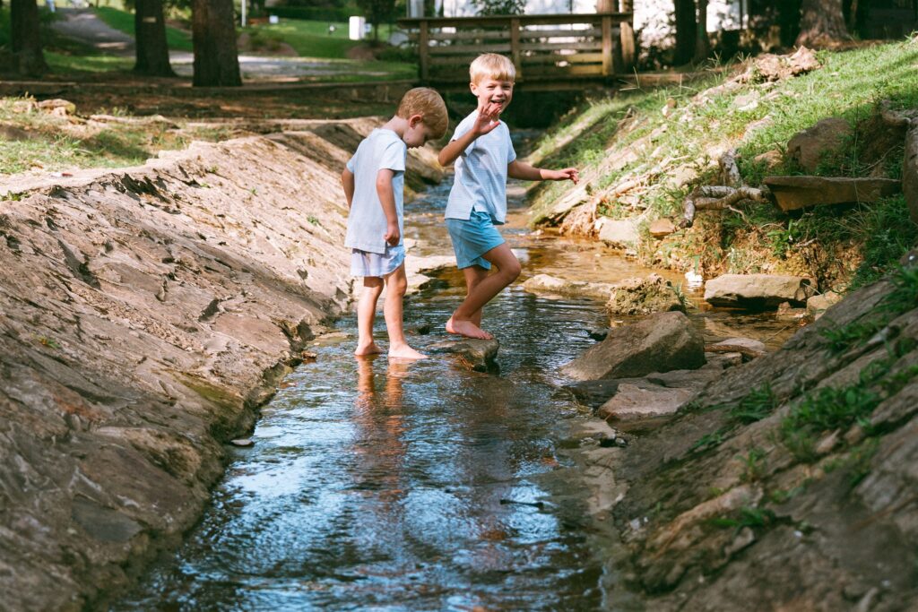 Two young boys wade barefoot through a creek during family photos at a park with a Birmingham family photographer. 