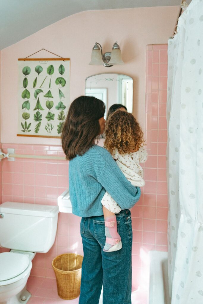 A mother & daughter stand before the mirror in their MCM pink bathroom during lifestyle family portraits in Birmingham AL.