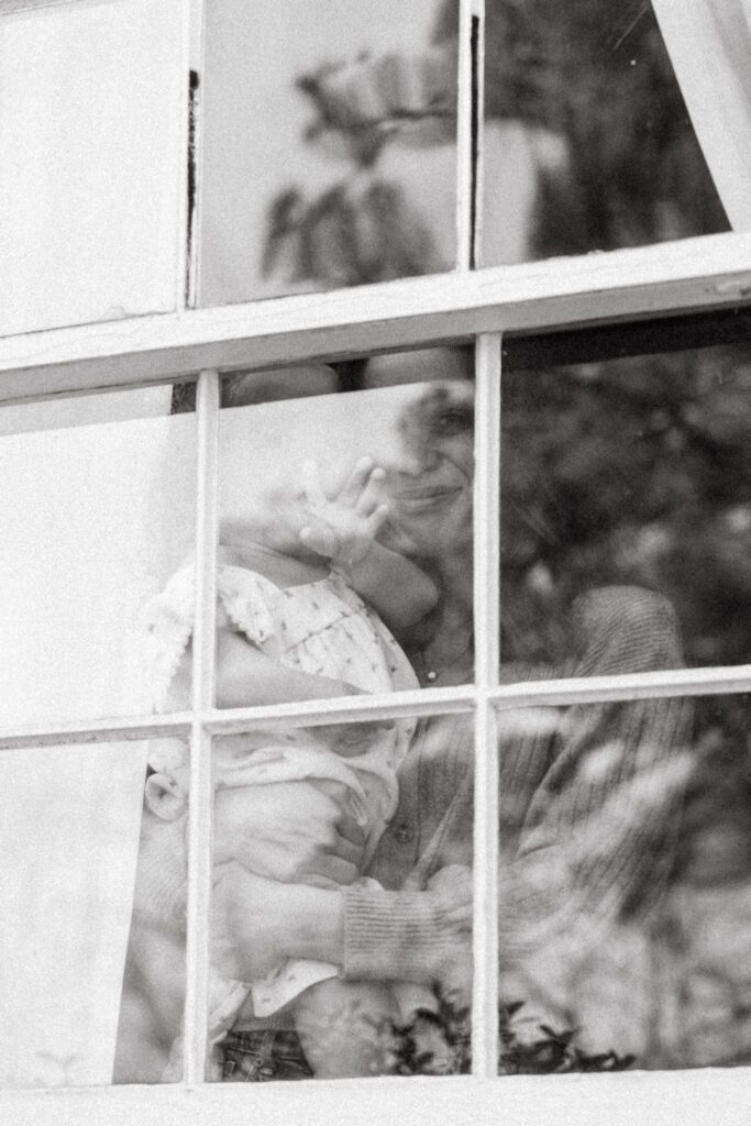 A b+w image depicts a mother holding her daughter through a paned glass window during an in home family photoshoot in Birmingham. 